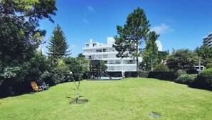 A grassy backyard with a small tree, a wooden deck, and a white building in the background.