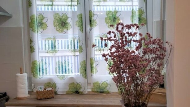Un bagno con una finestra ornata da tende floreali, un vaso con fiori secchi e un portascopevoli.