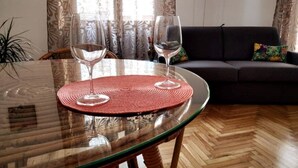 Un tavolo in vetro con due bicchieri da vino e un sottovaso intrecciato, un divano scuro e una tenda a motivi in un soggiorno.