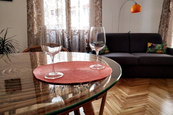 A glass table with two wine glasses and a woven placemat, a dark sofa, and a patterned curtain in a living room.