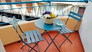 Un piccolo balcone con un tavolo blu e due sedie, un cesto di vimini con fiori e una vista su una barca sullo sfondo.