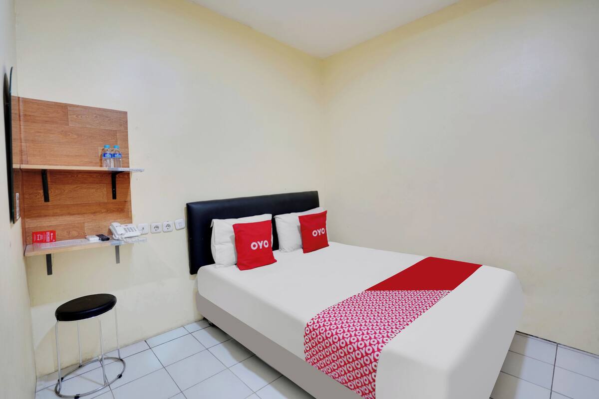 Habitación de hotel con una cama, un estante de madera con un teléfono y una botella de agua, y un taburete pequeño.