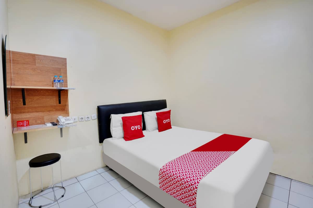Habitación de hotel con una cama, un estante de madera con un teléfono y una botella de agua, y un taburete pequeño.