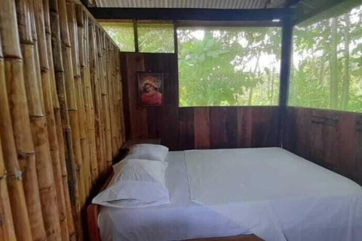Un lit simple avec du linge de lit blanc, dans une pièce aux murs en bambou et dotée d’une fenêtre donnant sur un paysage verdoyant.