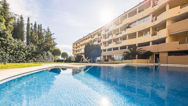 A swimming pool with a tiled deck, surrounded by a multi-story building with balconies and a garden area with trees and shrubs.