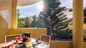 A balcony with a wooden table set for breakfast, including cups, a coffee pot, and a fruit bowl.