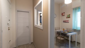 Un couloir mène à une salle à manger où une table est dressée pour deux personnes. On y trouve également une porte et une fenêtre avec des rideaux.