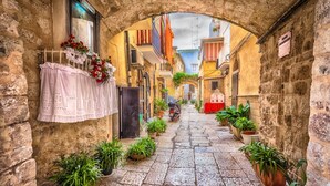 Une étroite ruelle pavée, ornée d’une arche rustique, de plantes en pot et d’un scooter garé au centre.