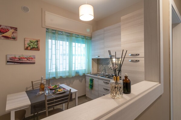 Cocina compacta con una mesa de comedor, una ventana con cortinas y una pequeña encimera con un florero y botellas.