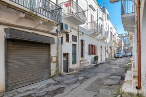 A narrow street with buildings on either side, closed shop shutters, and a clear blue sky.