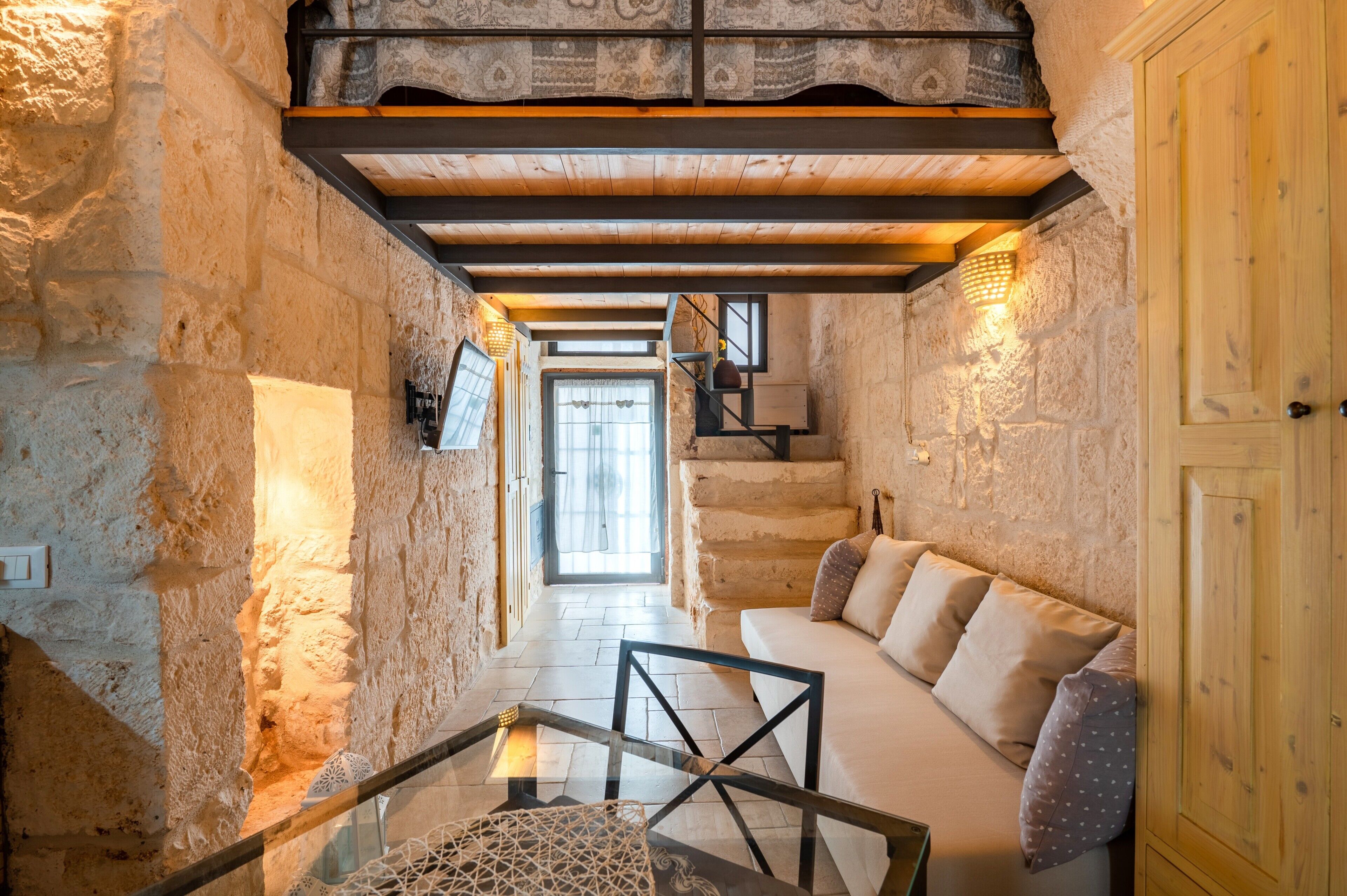 Ein Innenraum mit Steinsäulen, einer Holztreppe, einer Couch mit Kissen und einem Glastisch.