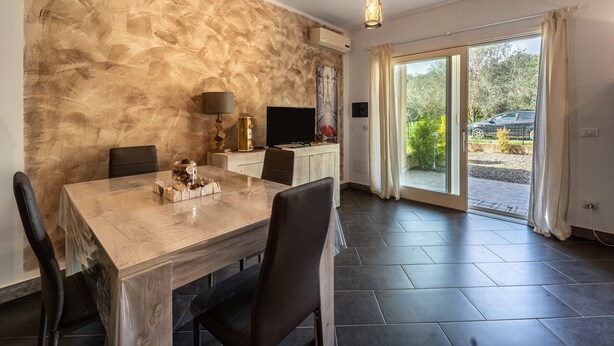 A dining area with a wooden table, chairs, a television, and a view of the outdoors through large glass doors. - Holiday Home "Open Space Nel Sulcis" with Private Garden & A/C (Masainas)