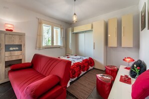 Une chambre avec un canapé rouge, un lit recouvert d’une couvre-lit à motifs floraux, une armoire en bois et un bureau avec une lampe rouge.