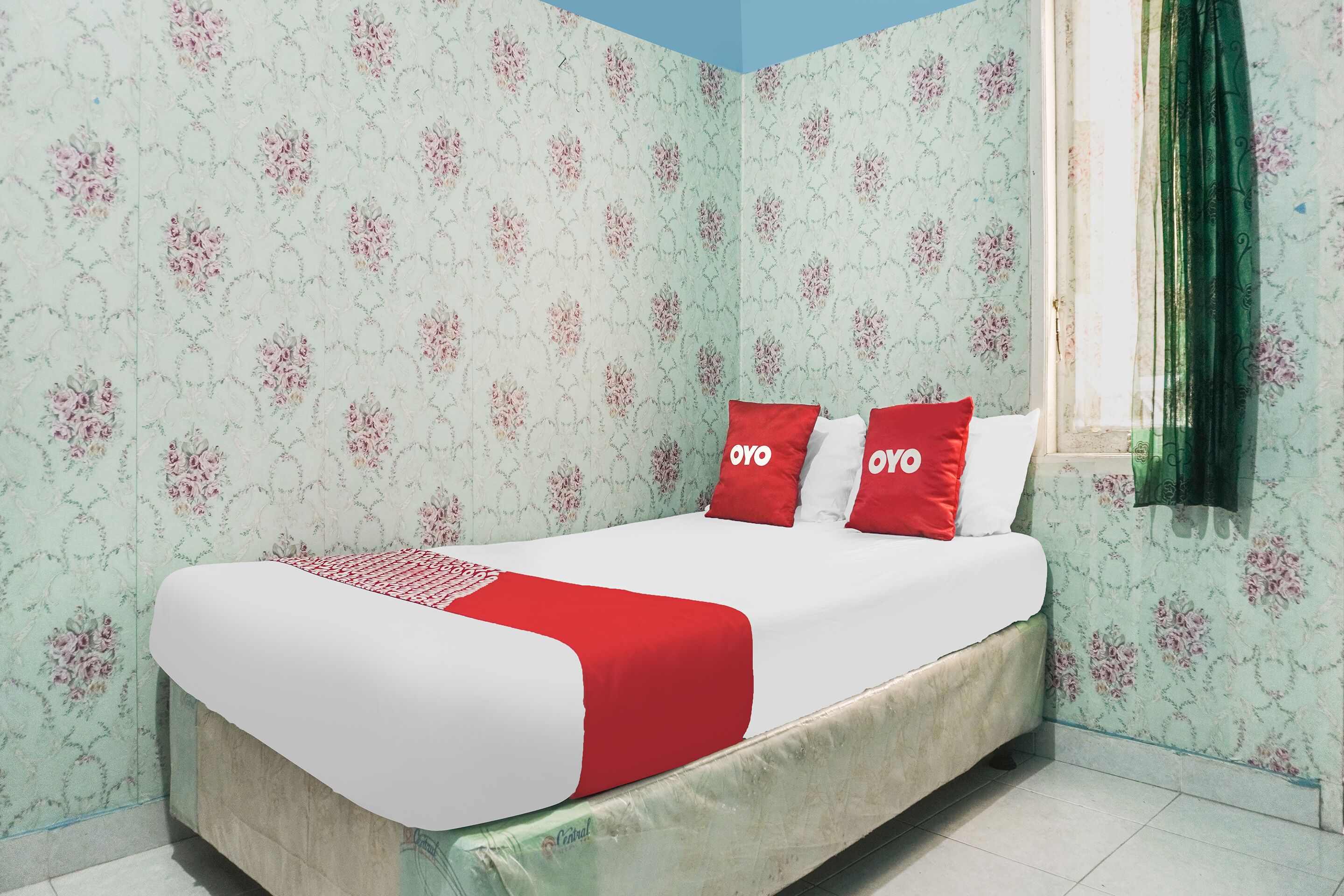A single bed with a red and white bedspread, two red pillows with "oyo" text, floral wallpaper, a window with green curtains, and a blue ceiling.