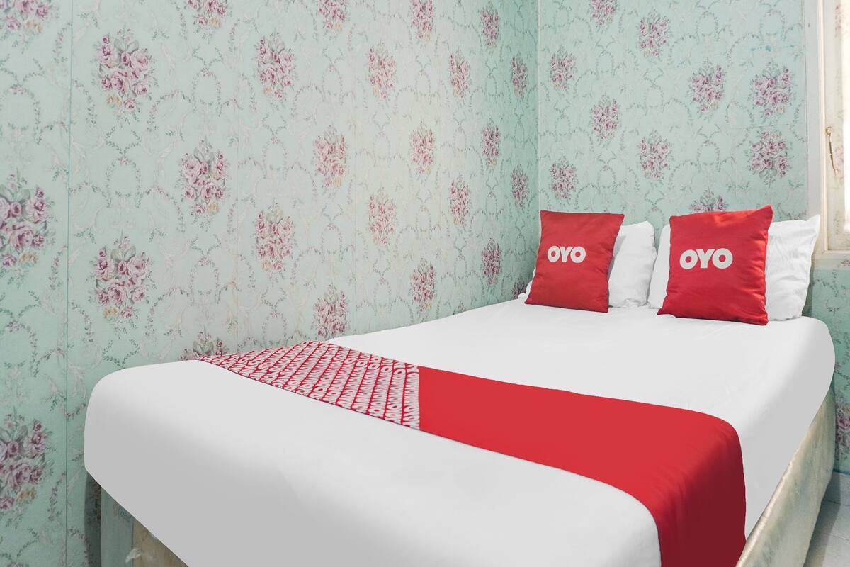 Una cama con ropa de cama blanca y roja, dos almohadas con el texto "OYO" y papel tapiz con motivos florales.
