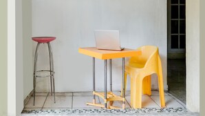 Une petite pièce avec un sol carrelé, un mur blanc, une fenêtre, une table avec un ordinateur portable, une chaise jaune et un tabouret rouge.
