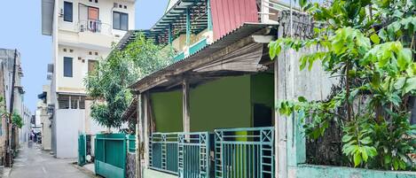 A narrow street with buildings on either side, including a two-story house with a balcony and a red roof.