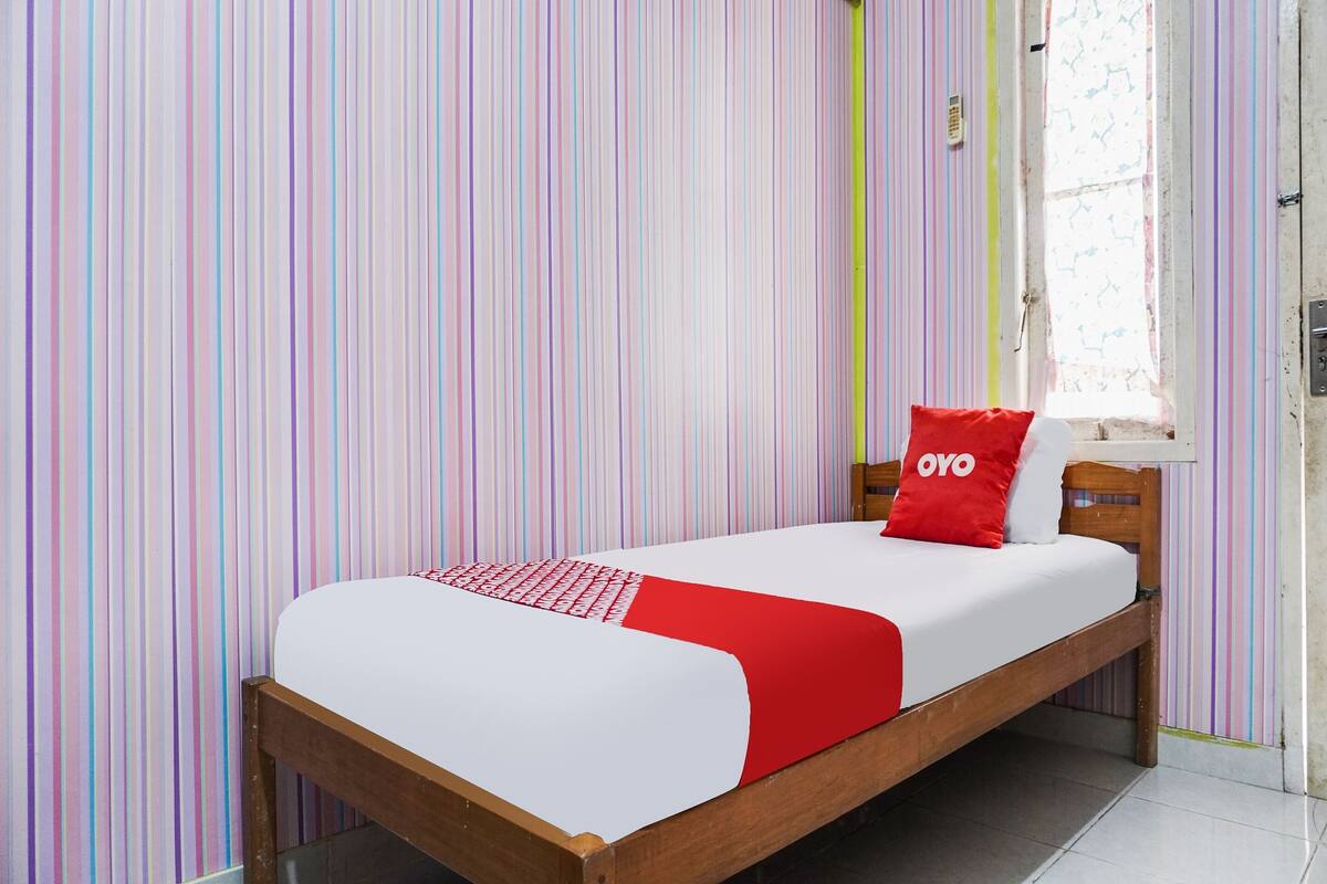 Una cama individual con una colcha blanca y roja, un almohadón rojo con el texto 'OYO' y un cabecero de madera.