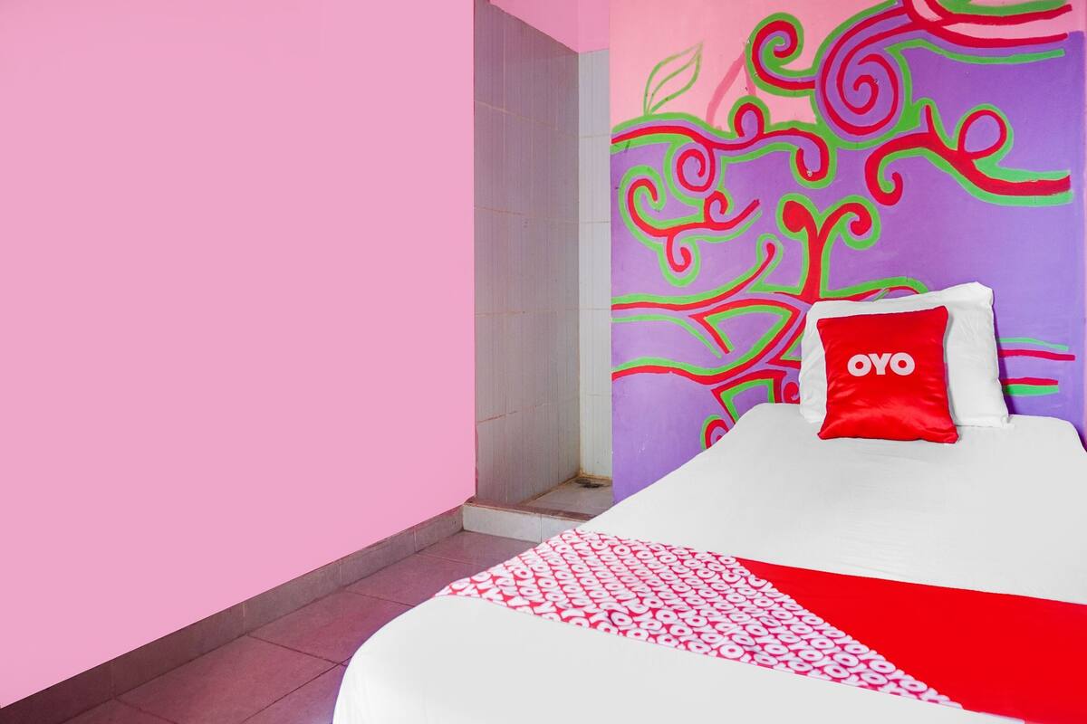 Habitación de hotel con cama, almohada roja con la marca 'OYO', ducha y pared con diseños abstractos de colores.