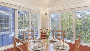 A dining area with a wooden table set for four, surrounded by wooden chairs, large windows with a view of trees, and a vase with white flowers. - 3875 The Hideout - Quiet Area, Walk to Town (Carmel-by-the-Sea)