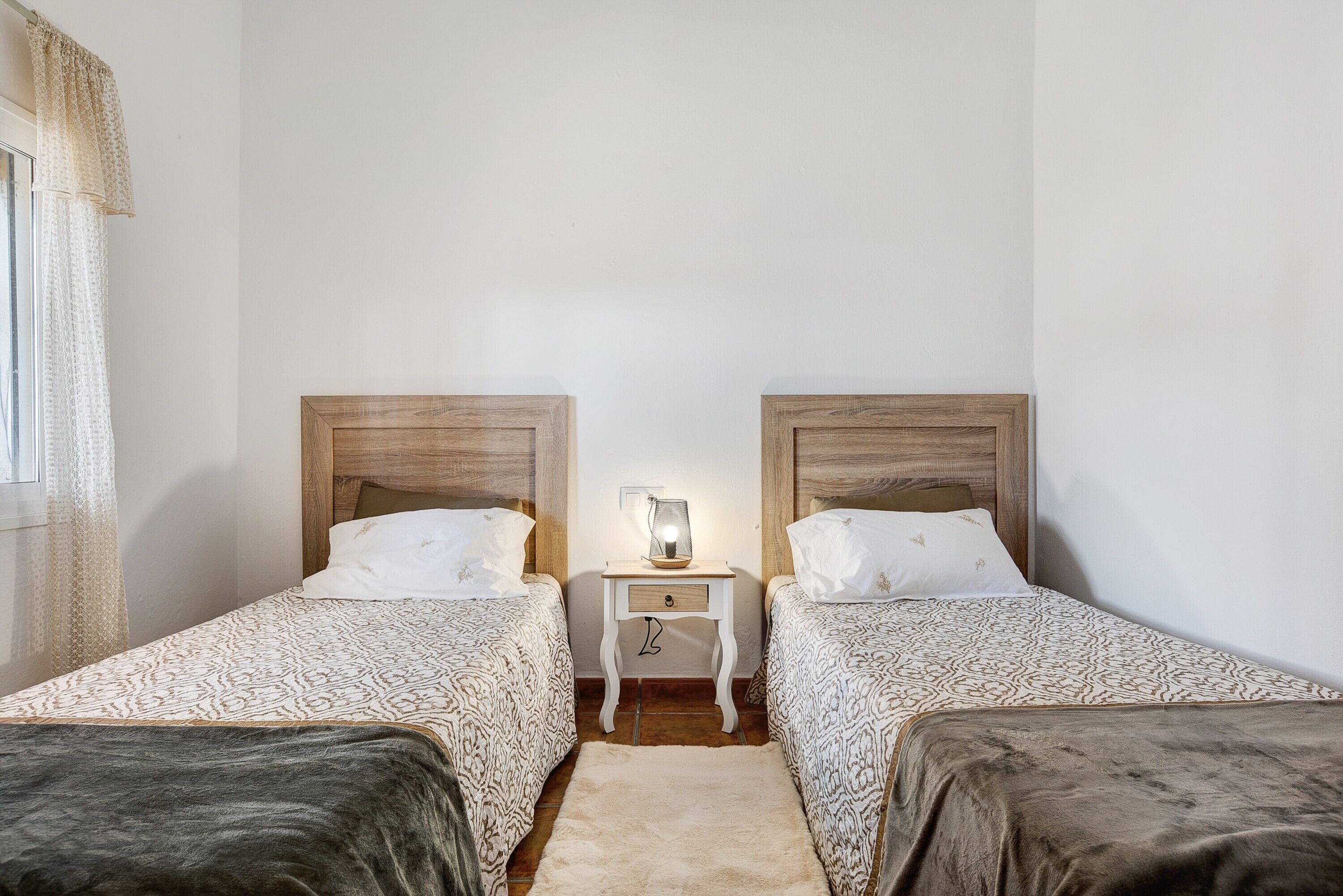 Una habitación con dos camas, una mesita de noche con lámpara y un ventanal con cortinas.