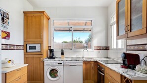 A kitchen with wooden cabinets, a washing machine, a microwave, a coffee maker, and a window with a view.