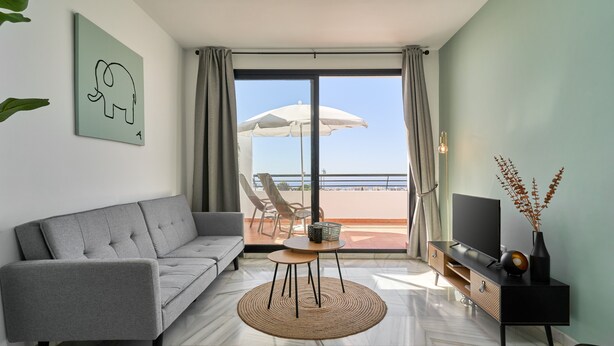 A modern living room with a grey sofa, a small round table, a TV on a black stand, and a view of the beach through a large window. - '2-Bedroom Apartment with Seaview', Wi-Fi and Air Conditioning (Nerja)