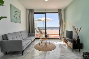 A modern living room with a grey sofa, a small round table, a TV on a black stand, and a view of the beach through a large window. - '2-Bedroom Apartment with Seaview', Wi-Fi and Air Conditioning (Nerja)