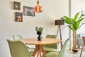 Ein Essbereich mit einem runden Holztisch, grünen Stühlen, einer hängenden Kupfer-Pendelleuchte, gerahmten Tierbildern an der Wand und einem Blumentopf.