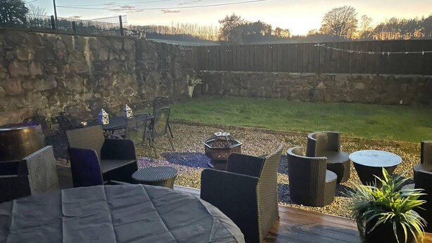 An outdoor patio area with a stone wall, a grassy yard, and a wooden fence. - Rural Chapel Cottage, Near Airport, bar & hot tub (Aberdeen)