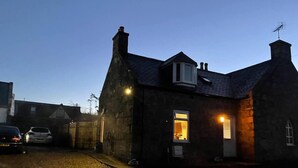 A stone house with a small porch and a single lit window, surrounded by a gravel driveway and parked cars. - Rural Chapel Cottage, Near Airport, bar & hot tub (Aberdeen)