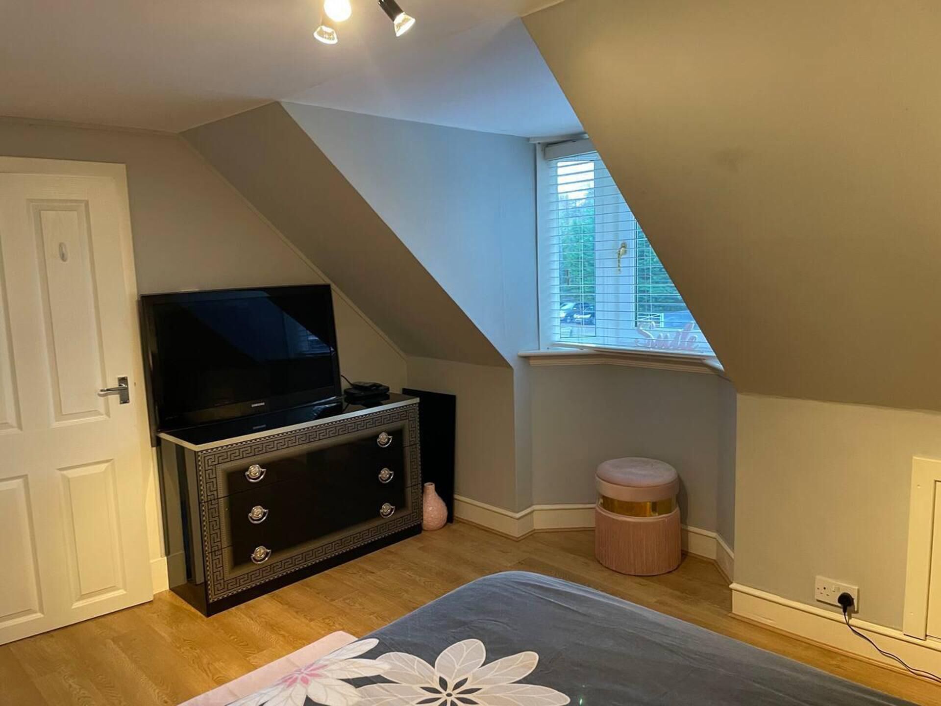 A bedroom with a TV on a dresser, a window with blinds, and a small round stool.