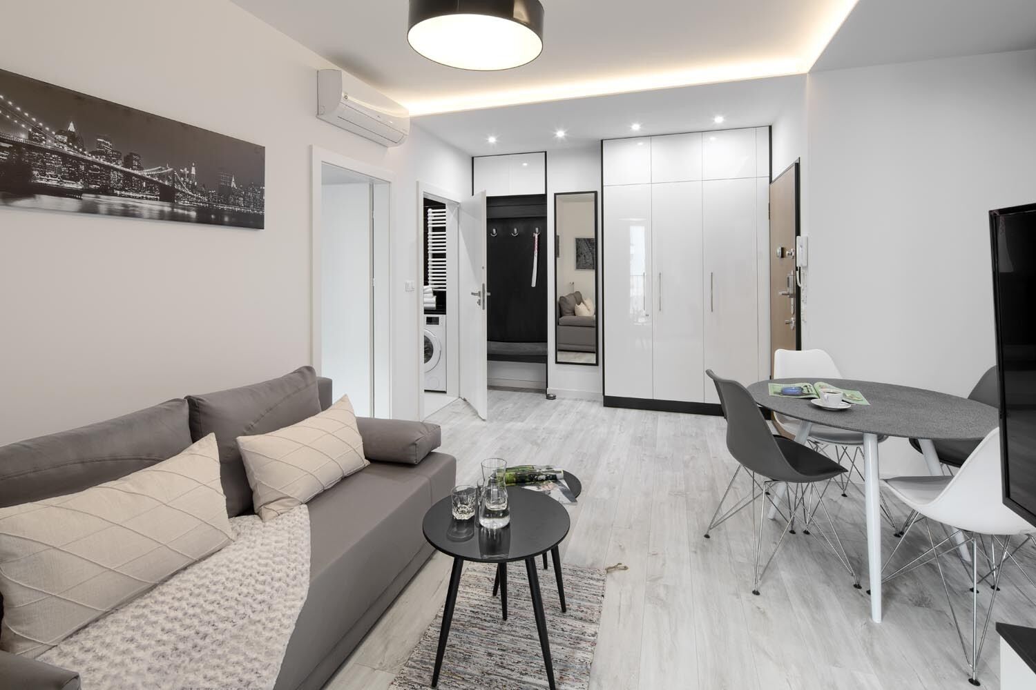 A modern living room with a grey sofa, a coffee table, and a dining area with a table and chairs. There is a cityscape painting on the wall, and a hallway leading to a bathroom with a mirror and a towel rack.