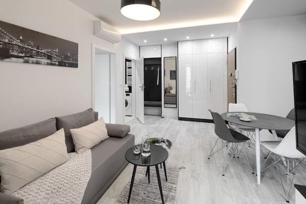 A modern living room with a grey sofa, a coffee table, and a dining area with a table and chairs. There is a cityscape painting on the wall, and a hallway leading to a bathroom with a mirror and a towel rack.