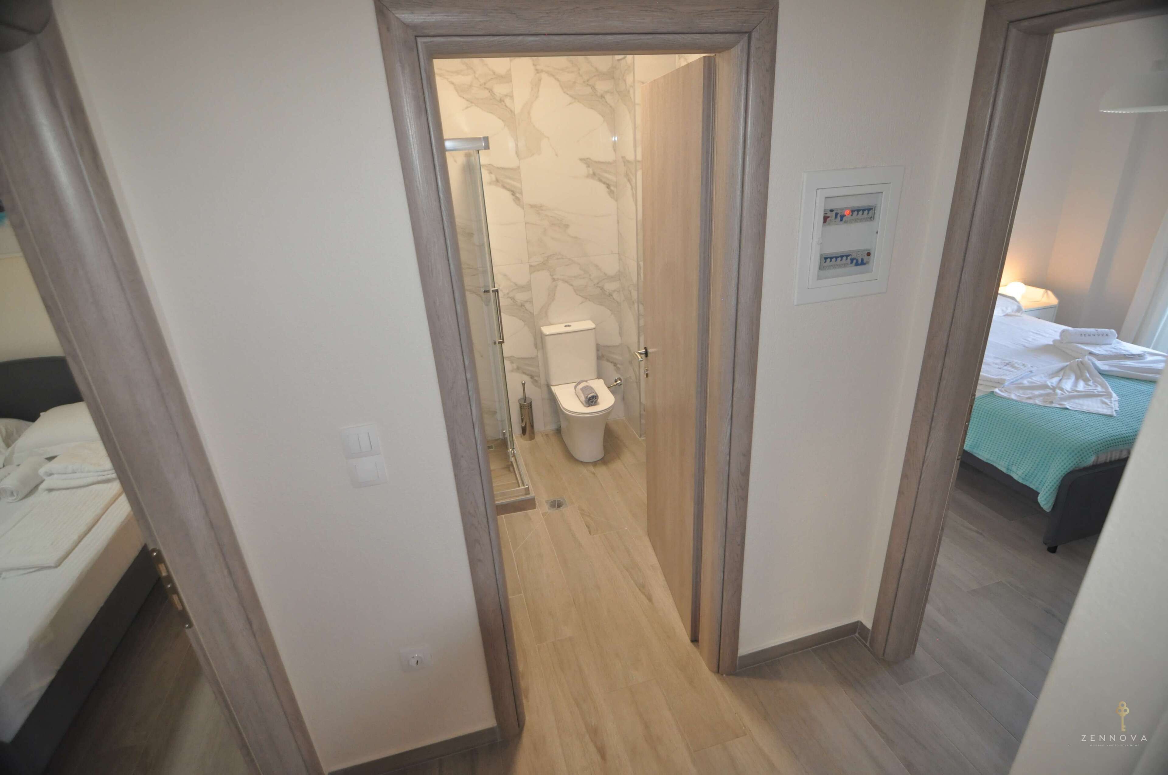 A narrow hallway leading to a bathroom with a toilet and a shower. There is a bed with a green bedspread in the adjacent room.