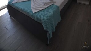A neatly made bed with white linens and a teal blanket, a single artwork on the wall, and a ceiling light fixture.