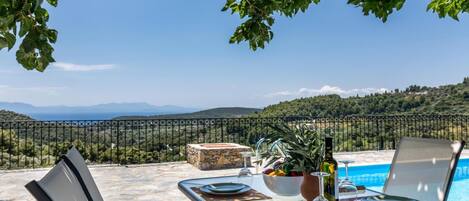An outdoor dining area with a view of the sea and mountains, a pool, and a stone table set for a meal.
