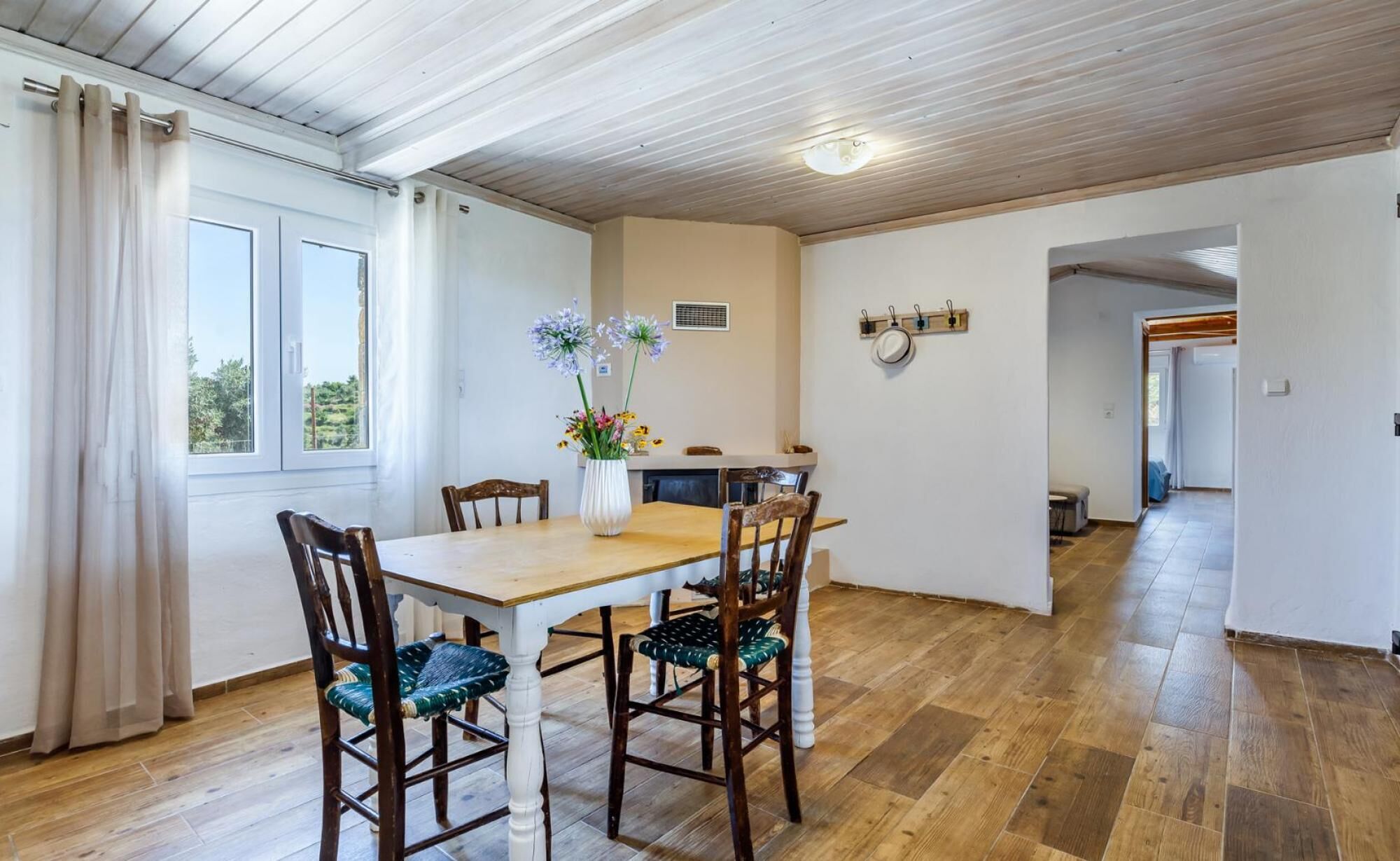 A dining area with a wooden table and chairs, a vase with flowers, and a view through a window.
