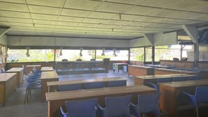 Une salle de classe avec des pupitres en bois et des chaises bleues, de grandes fenêtres et un plafond haut.