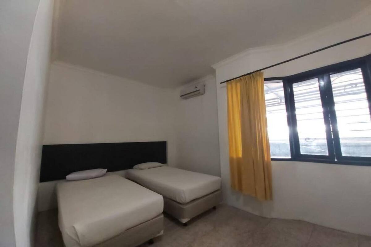 Une petite chambre simple, avec deux lits simples, une tête de lit, un appareil de climatisation et une fenêtre avec des rideaux.