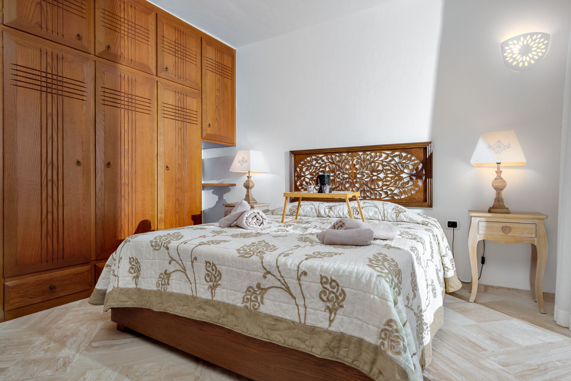 Een slaapkamer met een grote houten garderobe, een bed met een bloemenbedekking, twee bedlampjes en een decoratief hoofdeinde.