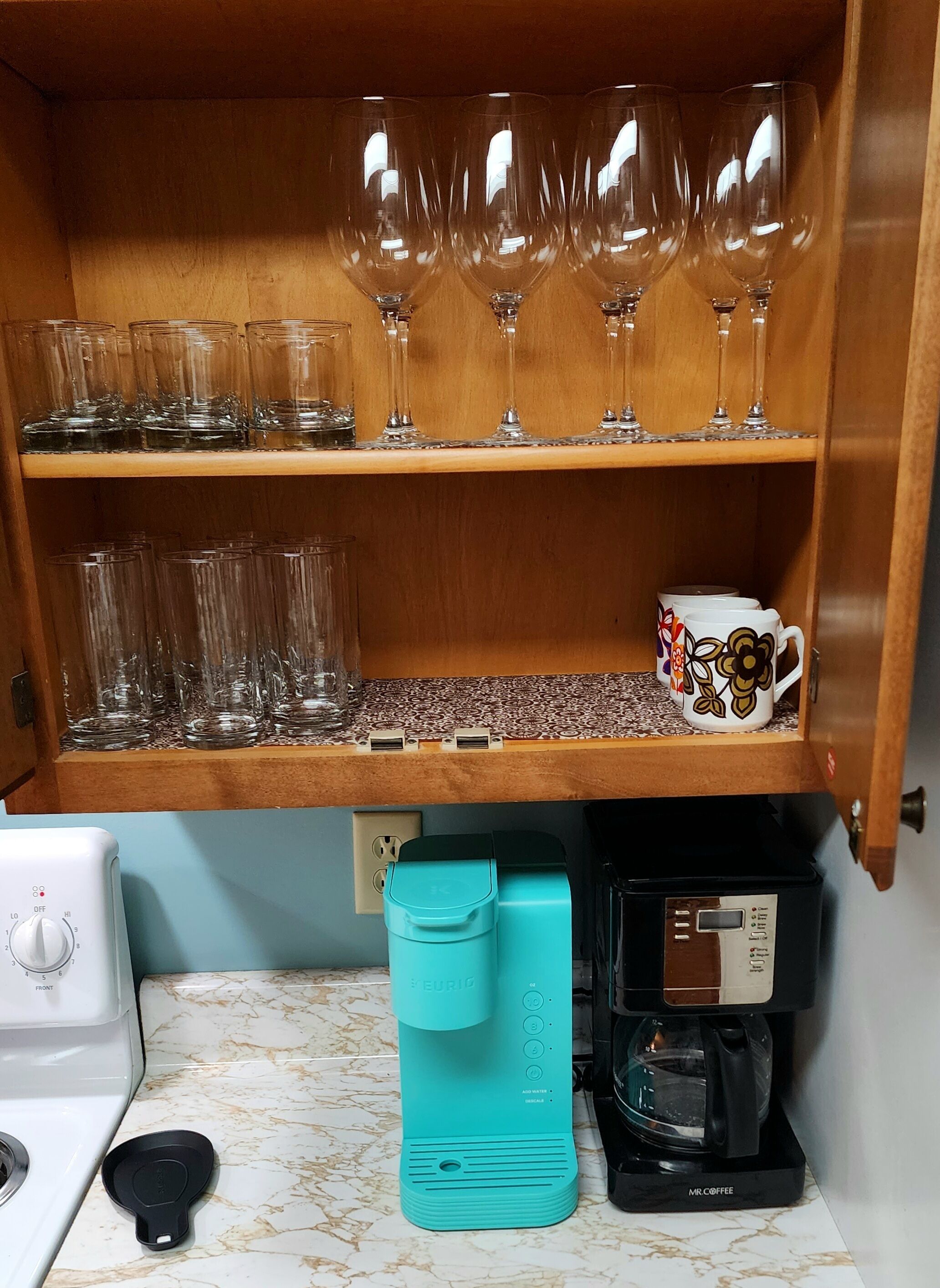 Una cocina con cafetera, hervidor y un armario con copas de vino y tazas.