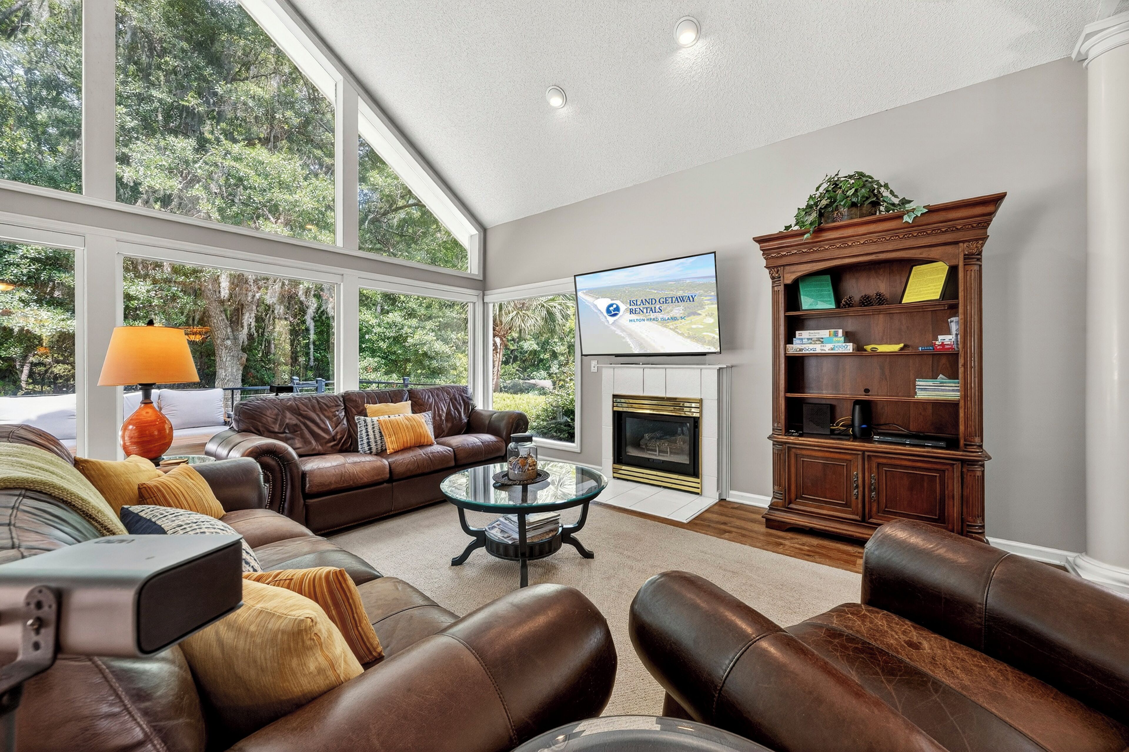 A living room with leather furniture, a fireplace, a television, and large windows overlooking greenery.