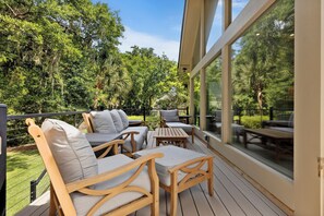 Una terraza de madera con mobiliario exterior, ventanas amplias y vistas a un paisaje verde exuberante.