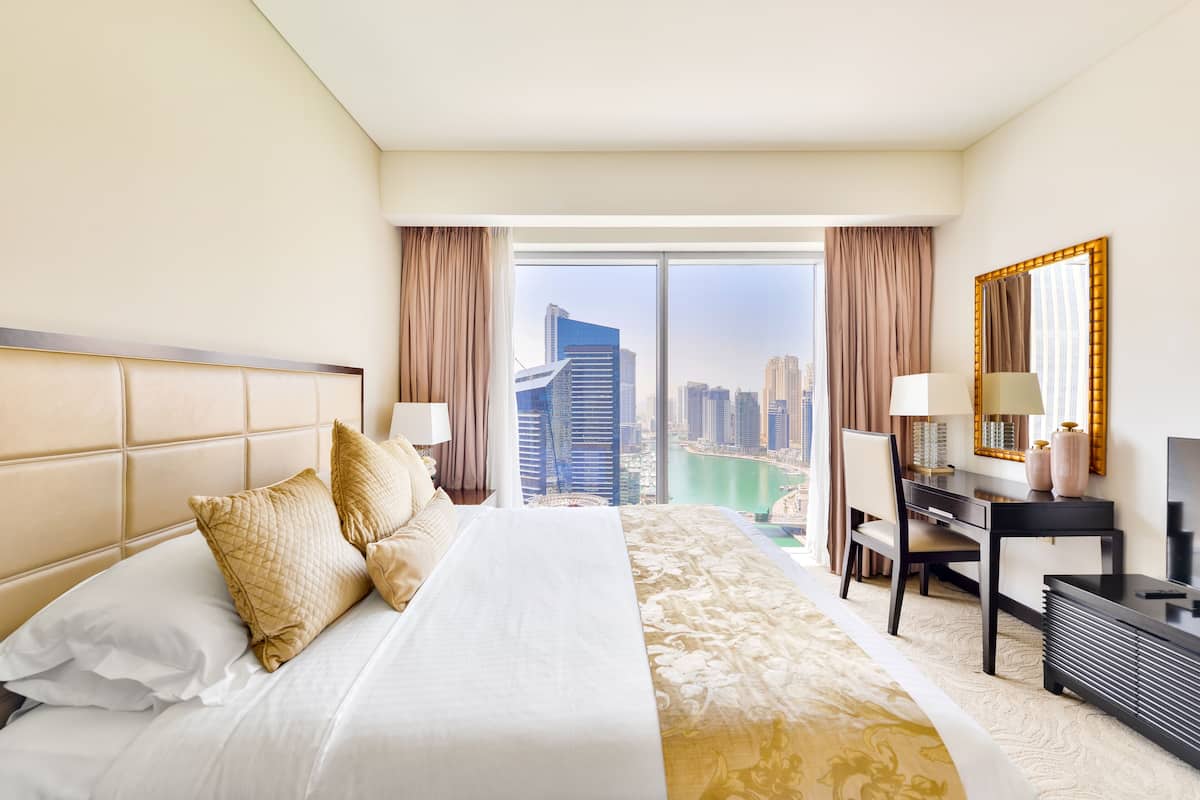 A hotel room with a large bed, a desk, a chair, and a view of a cityscape through a large window.
