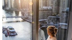 Una persona sentada junto a una ventana con vista a un río y barcos, en una habitación con cama y almohadas.