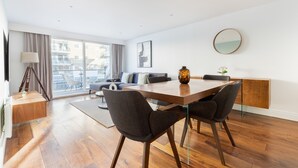 Un salon moderne avec une table à manger en bois, deux chaises grises, un canapé et une grande fenêtre avec des rideaux.