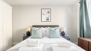 Une chambre à coucher bien aménagée, avec un grand lit, deux tables de chevet avec des lampes, une œuvre d’art encadrée au mur et une vue sur l’extérieur par une fenêtre ornée de rideaux.