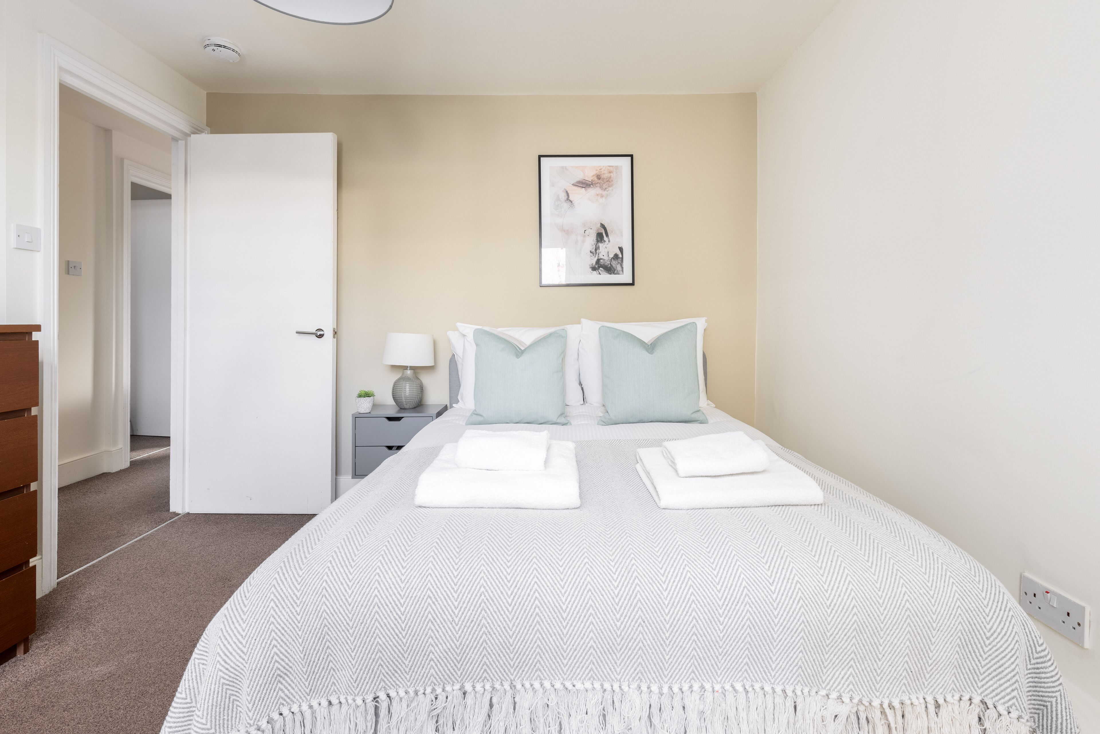 A bedroom with a bed, a bedside table with a lamp, and a door leading to another room.