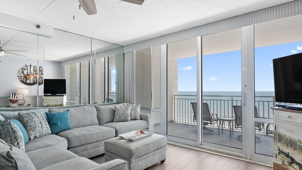 A modern living room with a grey sofa, a flat-screen TV, and a balcony with a view of the sea.